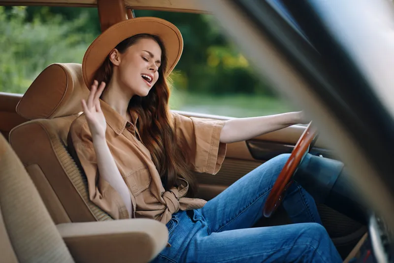 Happy young woman in a vintage car enjoying music, wearing a hat and casual outfit, surrounded by greenery, conveying a cheerful summer vibe.