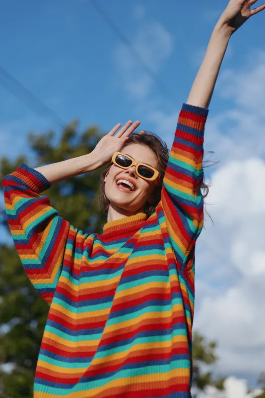 Woman in a rainbow sweater and sunglasses smiling, young adult portrait outdoors with arms raised, joyful fashion pose under blue sky, colorful casual outfit for lifestyle.