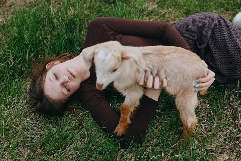 Young woman lying on grass outdoors embraces a gentle goat kid with affection, creating a warm lifestyle scene of bond between human and animal, serene playful mood, natural light, countryside vibe