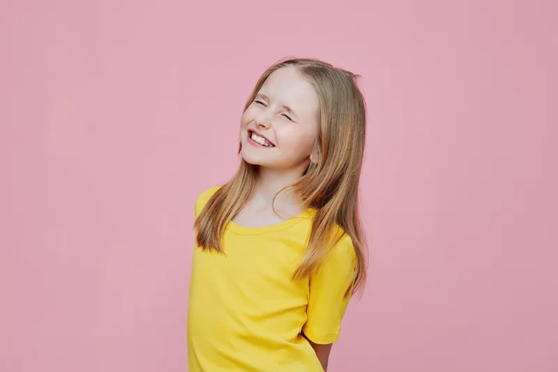 Laughing girl with long hair in a yellow shirt against a pink background, showcasing joy and positivity, perfect for children s marketing or lifestyle concepts