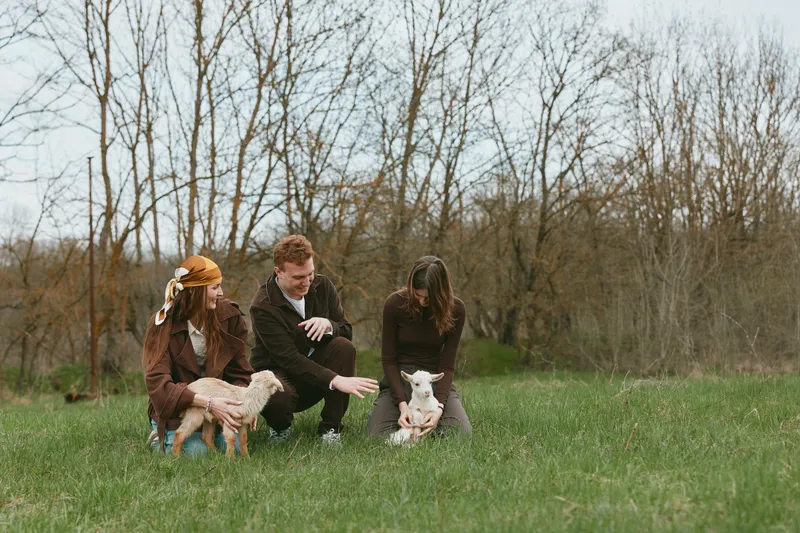 A family outdoors in a grassy field with a playful goat kid, an adult kneels smiling while children pet the animal, illustrating a wholesome rustic lifestyle and calm rural connection.