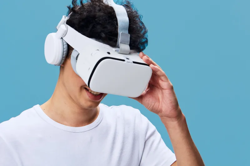 young man wearing virtual reality headset and wireless headphones enjoying immersive technology against pastel blue background, smiling and engaged in digital experience