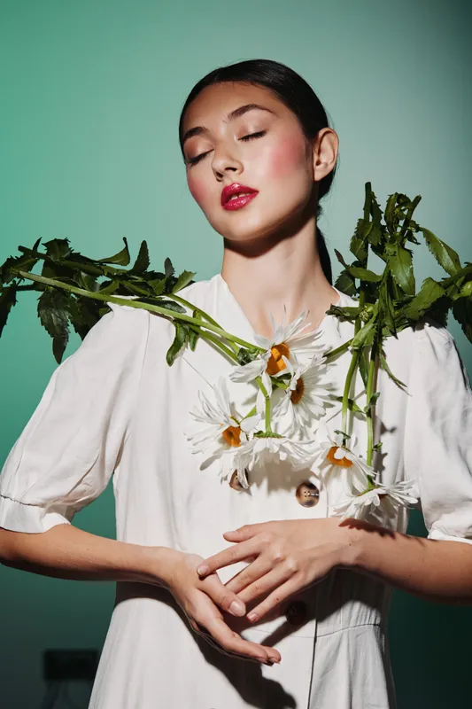 Young woman with bright red lipstick poses with closed eyes wearing white dress and holding large daisies against green background in creative beauty fashion concept.