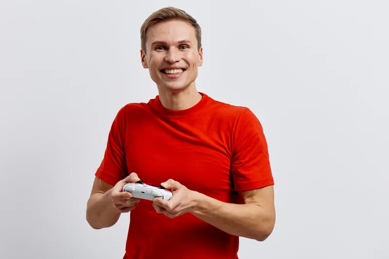 Smiling man holding a game controller, wearing casual red t-shirt, looking happy with excitement, isolated on plain white background. People lifestyle concept, entertainment, leisure, fun, gaming.