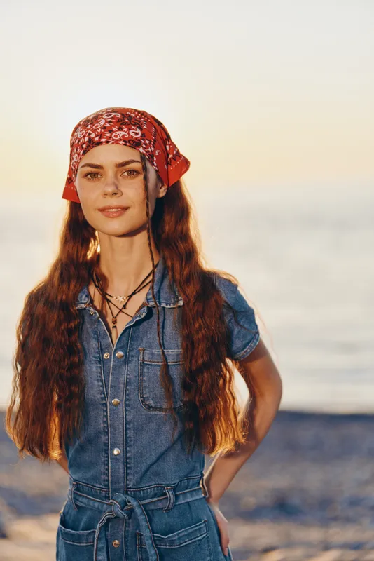 Lifestyle woman in modern boho-western denim street style with red bandana and layered necklaces, captured in warm film color tones at sunset by the sea.