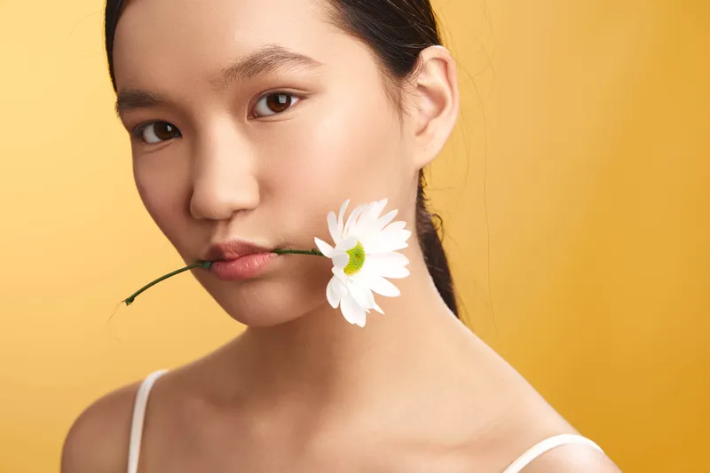 asian woman holds a delicate flower between her lips in a close up portrait, soft lighting and warm backdrop create a serene and elegant mood.