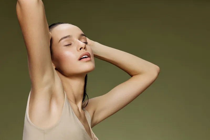Beautiful model with closed eyes poses with hands on head, showcasing natural beauty against a soft green background, emphasizing wellness and serenity