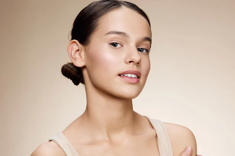 Young woman with a natural look, wearing neutral clothing, smiling gently against a soft beige background, conveying beauty and confidence through a flawless complexion
