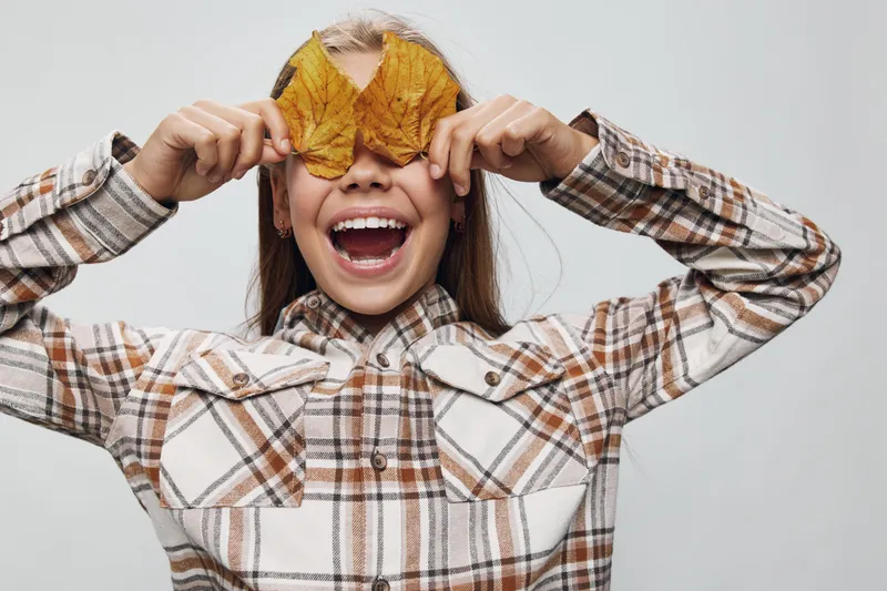 Child holding autumn leaves covering eyes and smiling with open mouth in plaid shirt. Cute kid having fun indoors with seasonal leaves and happy facial expression on light background.