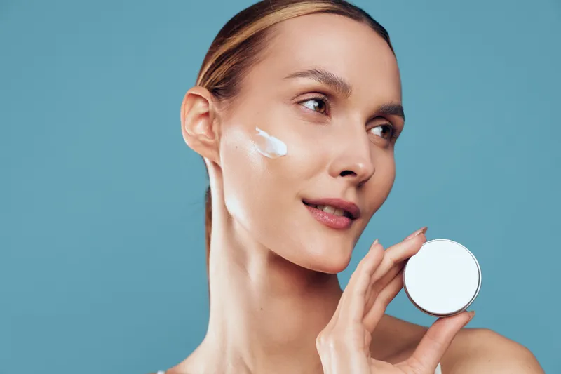 A woman applies cream to her cheek in a bright studio, highlighting skincare, hydration, and self care. Close-up reveals moisturizer texture and a simple daily routine for healthy skin.