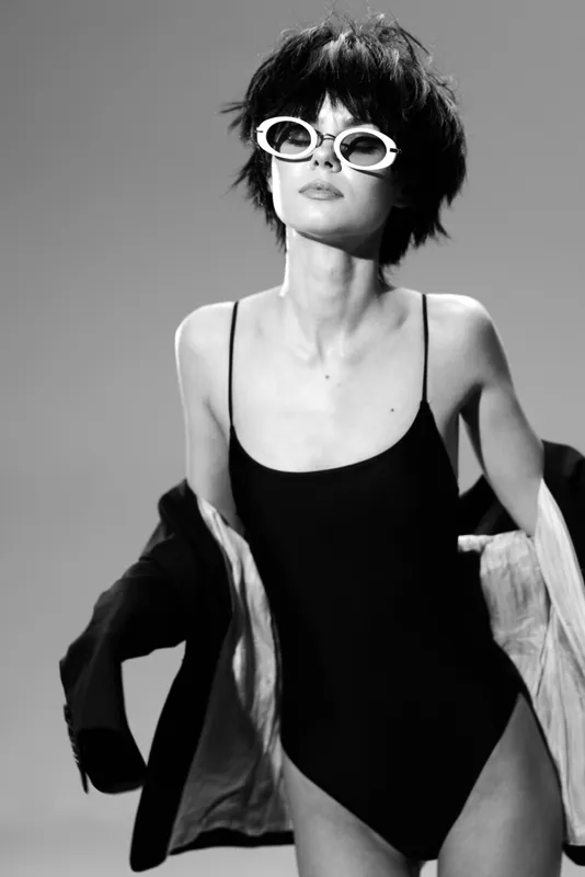 Black and white portrait of a stylish woman wearing sunglasses and a black swimsuit with a jacket, posing confidently in studio lighting.