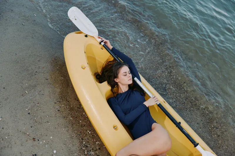 A woman rests inside a yellow kayak on a pebbled shoreline, holding a paddle and gazing toward the water, capturing calm outdoor adventure and coastal relaxation.