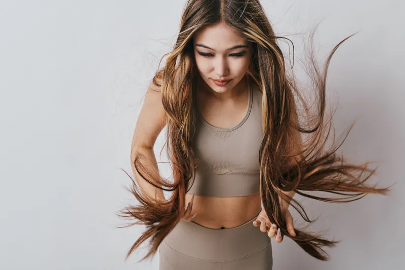 Young woman in sportswear with long flowing hair looking down, fitness concept, indoor studio with neutral background and natural light for active lifestyle.