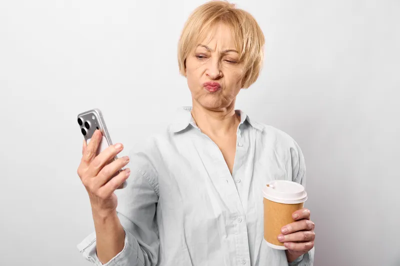 Woman smartphone coffee senior indoors, face communication beverage, holding a coffee cup, looking at the screen with focused expression, standing in background, posture, comfort