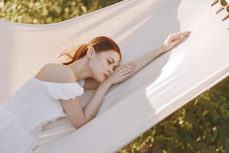 Young woman relaxing in a hammock outdoors, wearing a white summer dress, serene expression, enjoying nature surrounded by greenery during warm season