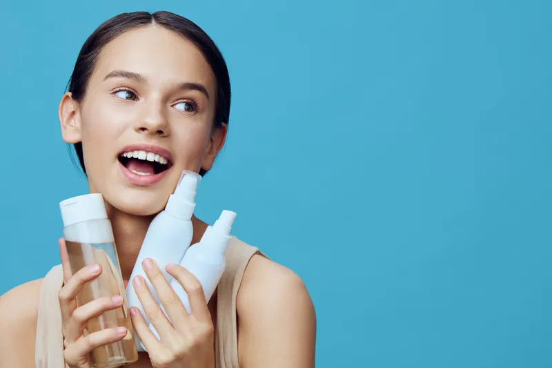 Young woman showcasing skincare products with a cheerful expression against a bright blue background, emphasizing healthy beauty and self care