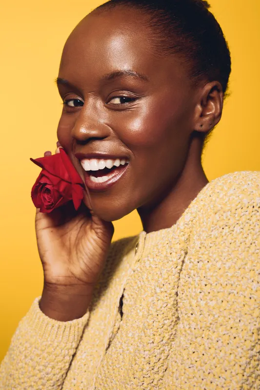 Smiling African woman with dark skin holds red rose near face. She wears a beige knit sweater on yellow background. Concept of beauty, happiness, natural skin, and floral aesthetics.