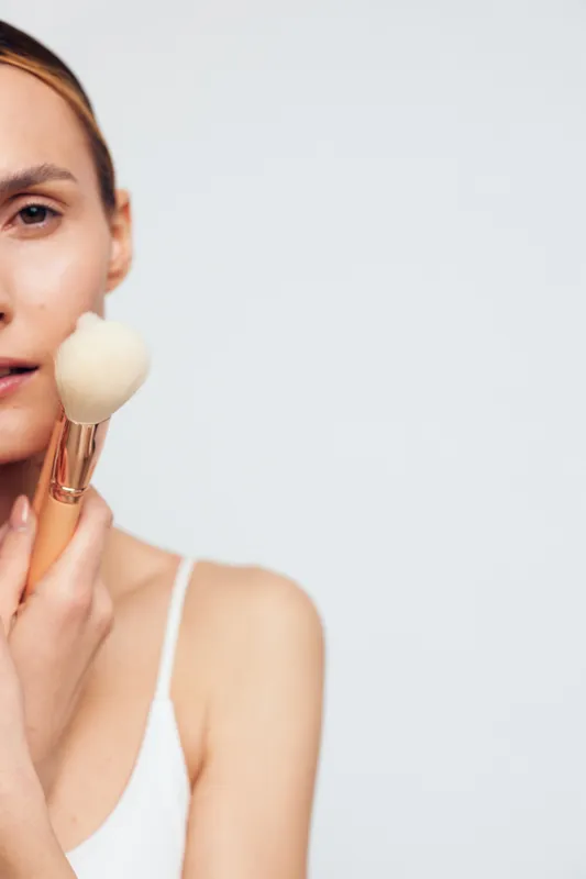 Close-up of a young woman applying makeup with a large soft brush on her face. Clean skin, natural look, and minimalistic background create a fresh beauty concept.
