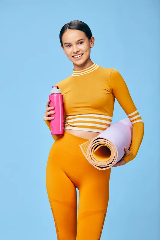 Fitness woman in bright orange athletic wear holds a pink water bottle and rolled yoga mat, ready for a dynamic workout and sport training session.