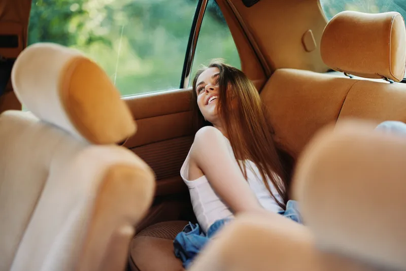 Young woman relaxing in the backseat of a car, enjoying the moment and sunshine, with a carefree expression and long hair, perfect for lifestyle content.