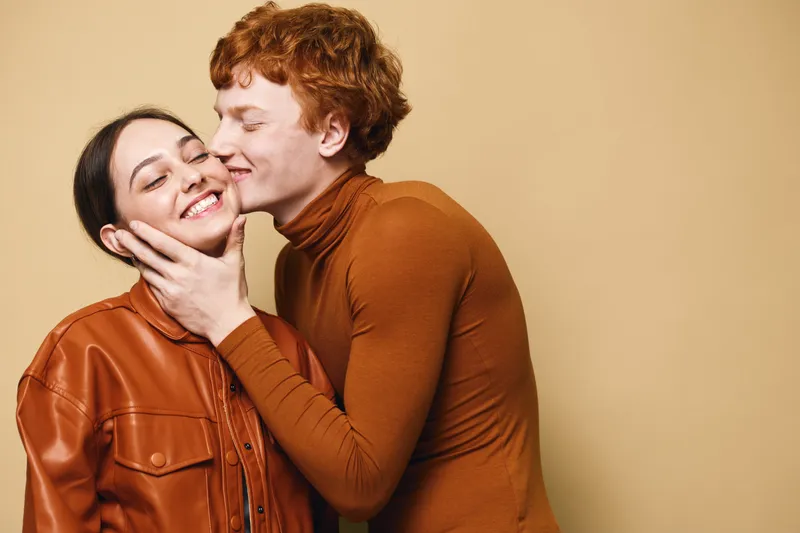 Young couple in warm brown clothing sharing a tender moment, smiling and embracing against beige background. Romantic gesture with affection and close connection between two people.
