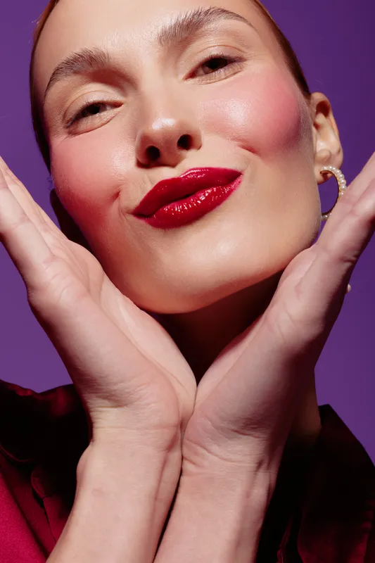 Close-up of a smiling woman with red lipstick and smooth skin posing with hands near face on purple background. Concept of beauty, makeup, skincare, and confident expression.