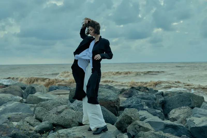 Windy coast photo of a woman in a dark coat and white dress posed on rocky shore, sea spray and cloudy sky create dramatic outdoor fashion mood.