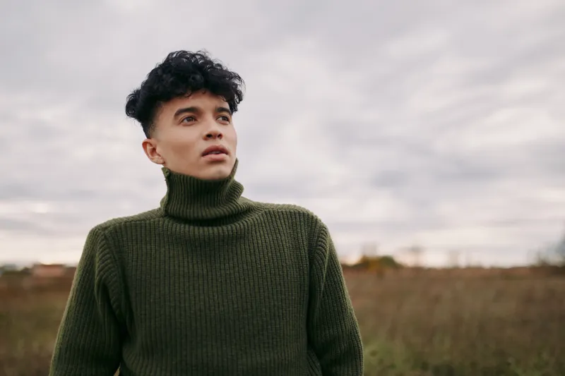 A young man in a green turtleneck stands in an open field, portrait captures an authentic, credible candid moment with natural expression and relaxed posture under cloudy sky.