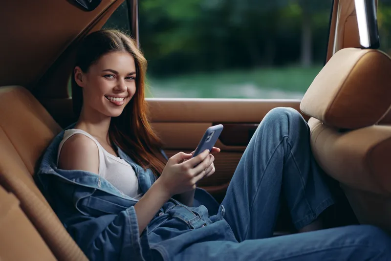 Young woman in casual denim outfit smiling while using smartphone in car interior, reflecting joy and comfort during spring or summer season.