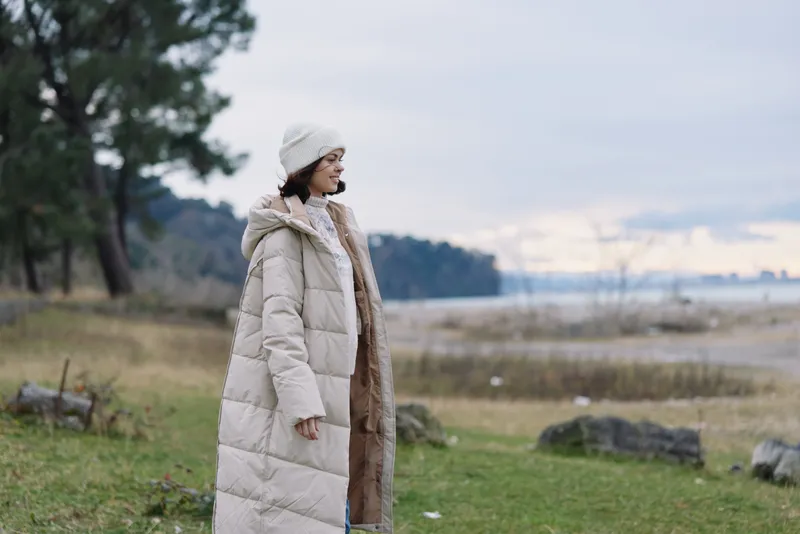 Woman in a long beige puffer coat and hat standing near a lake, autumn park landscape, calm sky, distant trees, soft light, outdoor fashion and travel mood.