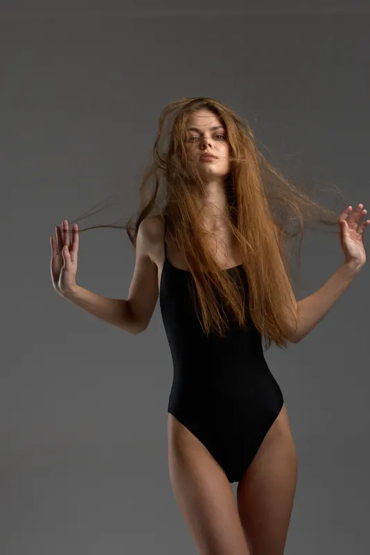 Young woman with long red hair wearing black bodysuit posing against gray background with serious expression.