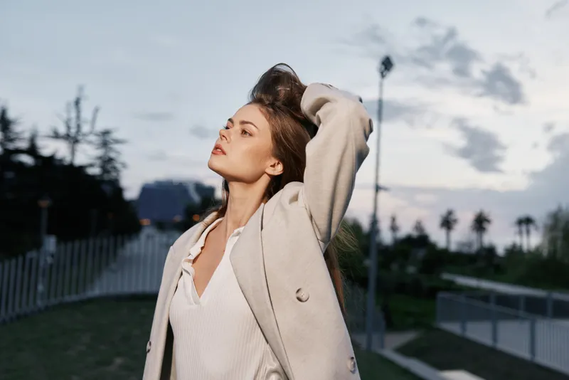 Young woman in beige coat outdoors with confident expression during cloudy sunset, natural light, autumn fashion concept