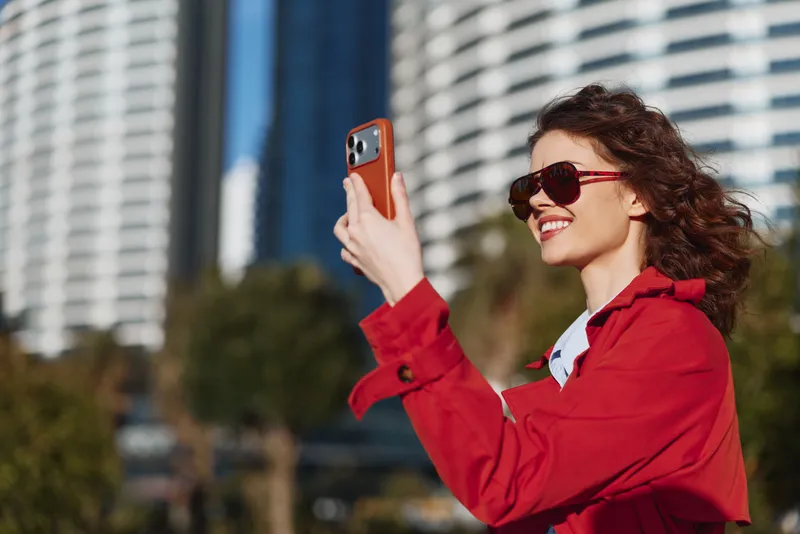 Young woman with curly hair wearing a red coat and sunglasses smiles while taking a selfie outdoors in an urban environment on a sunny day.