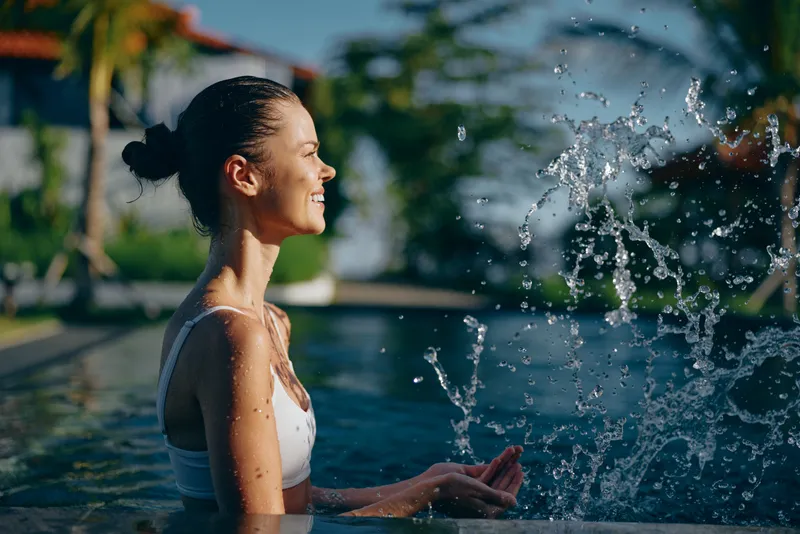 Young woman in white swimsuit enjoys sunny day in outdoor pool smiling with closed eyes and splashing water with hands in tropical lifestyle setting.