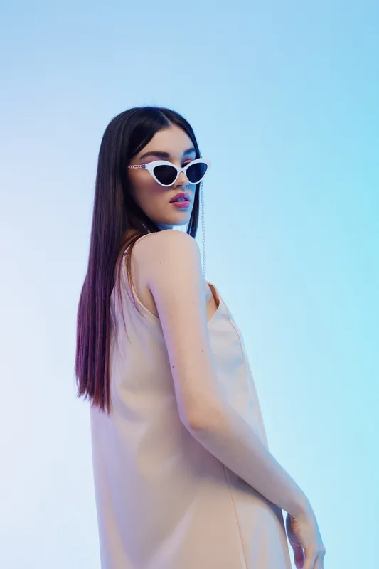 Stylish woman with long dark hair wearing white sunglasses and a beige dress poses against a gradient blue background. Concept of beauty fashion and elegance.