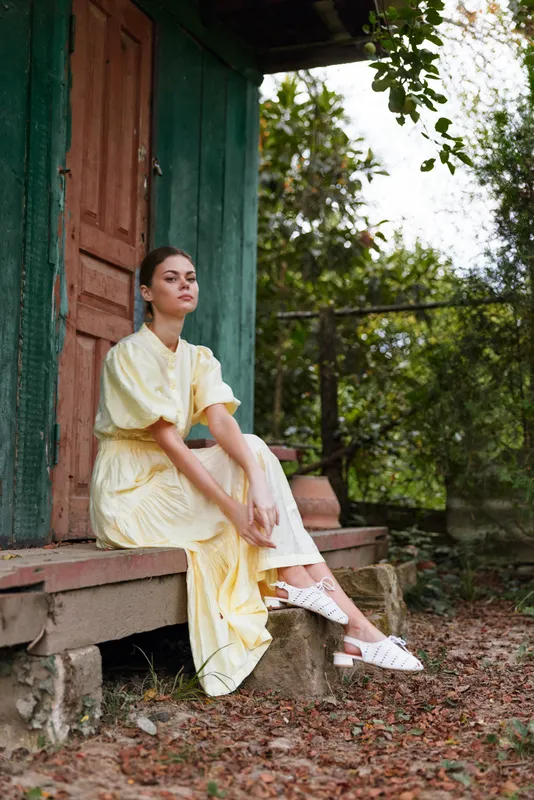 Woman in a pale yellow dress sits on a porch in a tranquil outdoor setting, smiling softly, relaxed pose, natural light and greenery around today