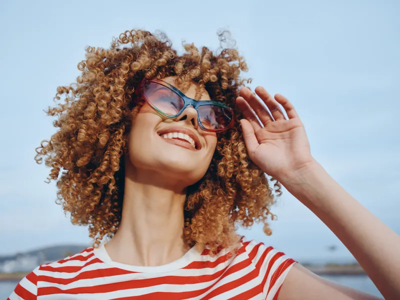 Portrait of a woman with voluminous curly hair, wearing a striped shirt and sunglasses, smiling brightly under a clear blue sky, capturing carefree summer energy and sunshine.