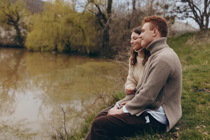 Cheerful couple outdoors, seated by a calm river, cozy sweaters, gentle breeze, relaxed posture, natural tones, soft light, lush greenery edge, tranquil moment, genuine companionship, scenic lifestyle