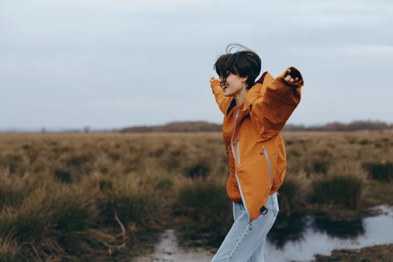 Woman lifestyle outdoors dog jacket walking nature lake countryside field background, woman in orange jacket smiles and guides small pet on leash, overcast sky, peaceful daily life