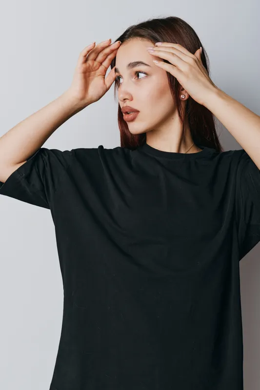 Young woman with long hair wearing black shirt touching forehead looking to side with thoughtful expression on gray background in studio portrait with natural makeup and earrings