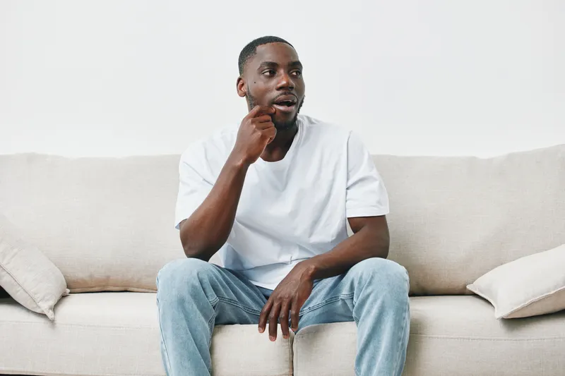 Thoughtful man sitting on a couch in a casual white t shirt and jeans, expressing contemplation in a modern, minimalistic living room setting