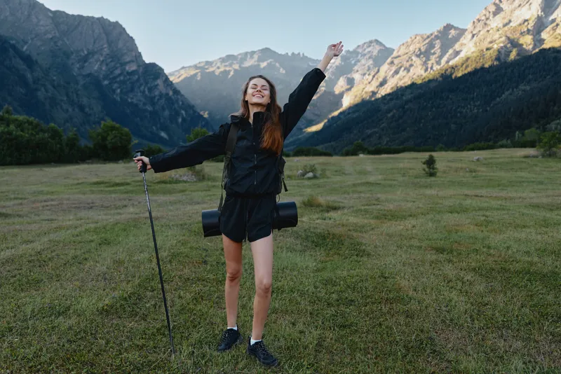 A cheerful female hiker stands in a wide mountain meadow, smiling with arms raised, embracing the outdoors and a sunny day, exuding energy and accomplishment.