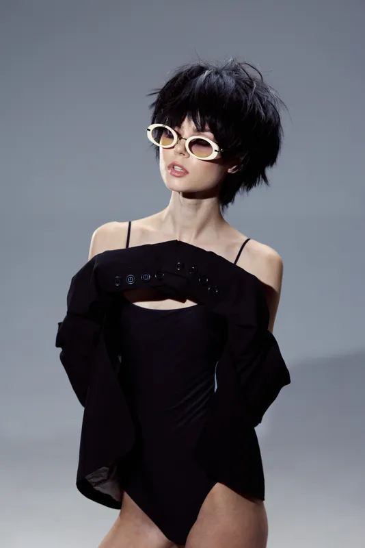 Stylish woman in black outfit wearing sunglasses with short hair posing against gray background