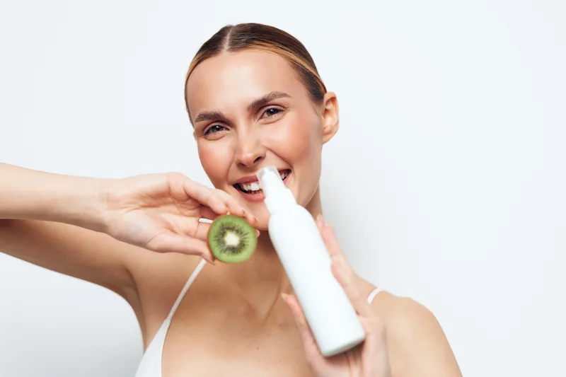 Young woman with blonde hair smiles joyfully while holding a white bottle and a sliced kiwi in front of a white background.
