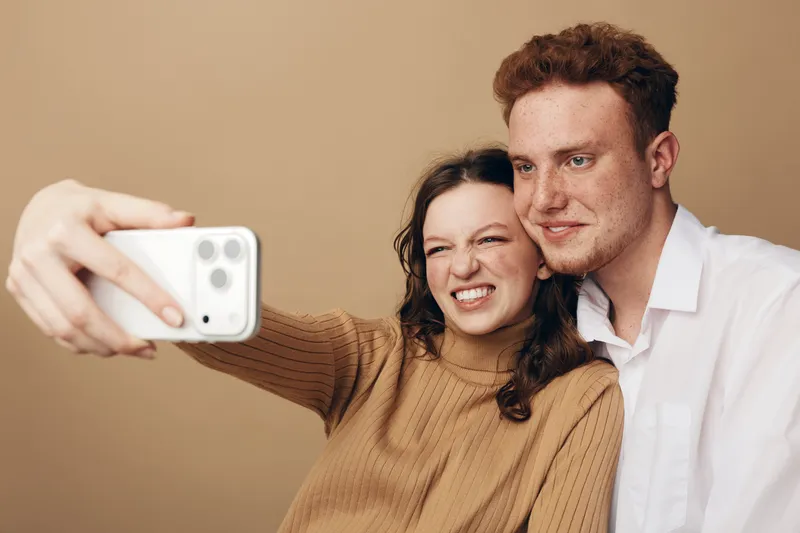 Young couple takes a selfie with a smartphone. Woman in brown sweater makes a funny face while man in white shirt smiles. Indoor casual moment with warm beige background.