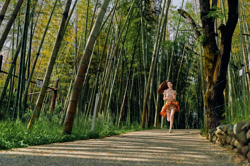 Woman running through a bamboo forest path in nature, trail lined with tall green trunks and sunlit leaves, motion captured as she moves toward camera in a flowing skirt, active summer scene.