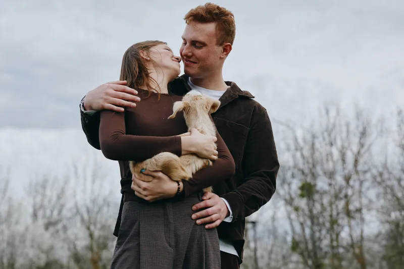 A loving couple holds a goat kid outdoors, sharing a gentle kiss and warm smiles. Friendly animal interaction creates a wholesome lifestyle moment in soft natural light.