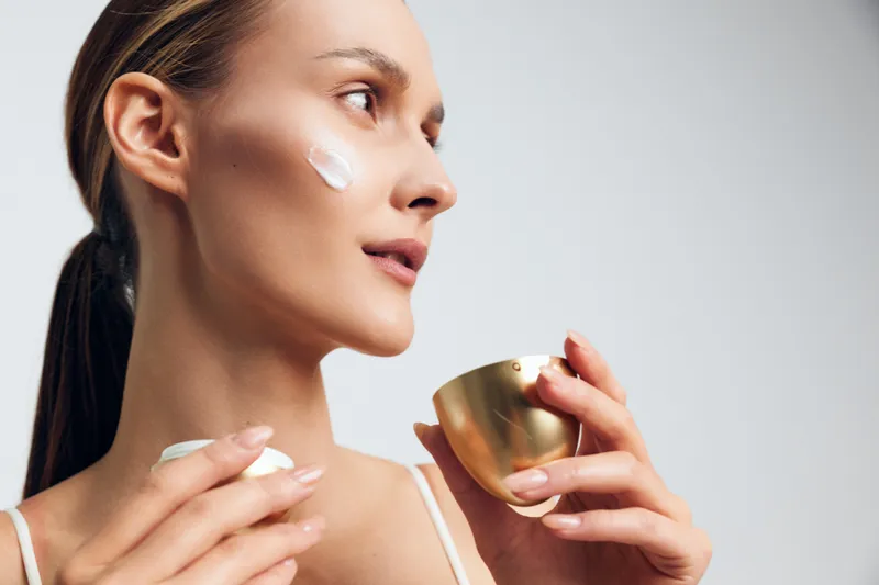 Young woman applying face cream with smooth skin, holding golden jar, natural skincare routine, soft lighting, clean background, beauty treatment concept.