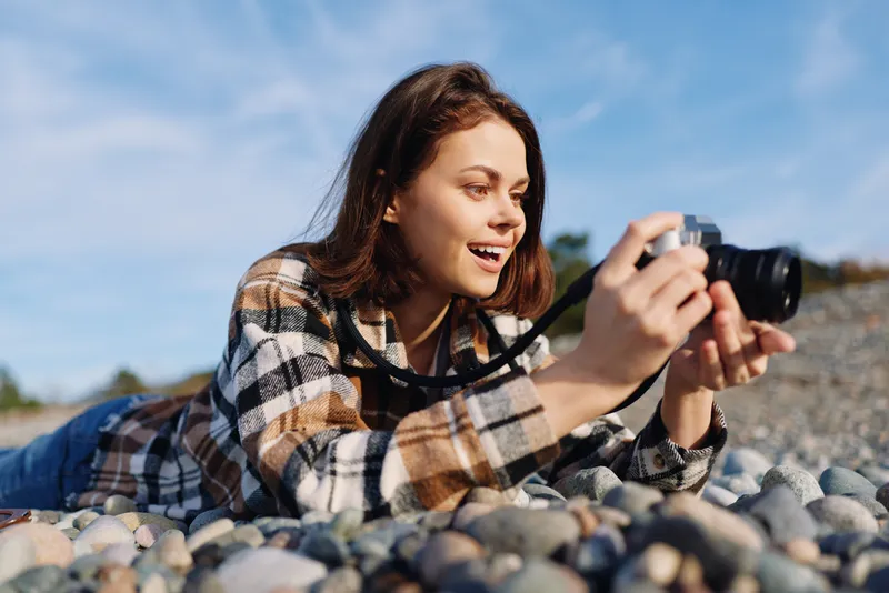 Woman on a rocky beach uses a camera for candid photography, wearing a plaid shirt, capturing focused moments in natural light with a relaxed outdoor vibe