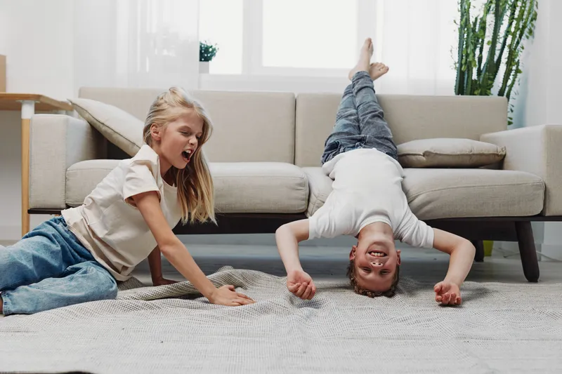 Two playful children enjoy fun time indoors, one girl laughing on the floor and boy doing a headstand on carpet near sofa, bright living room with modern decor.